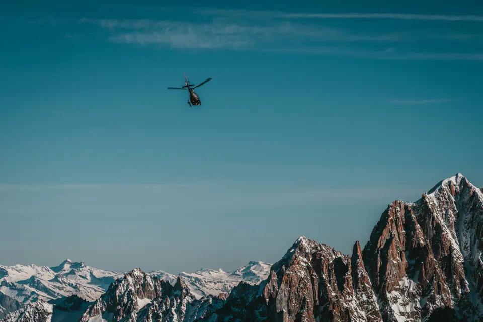 A black helicopter flying above jagged snow-capped peaks under a deep blue sky