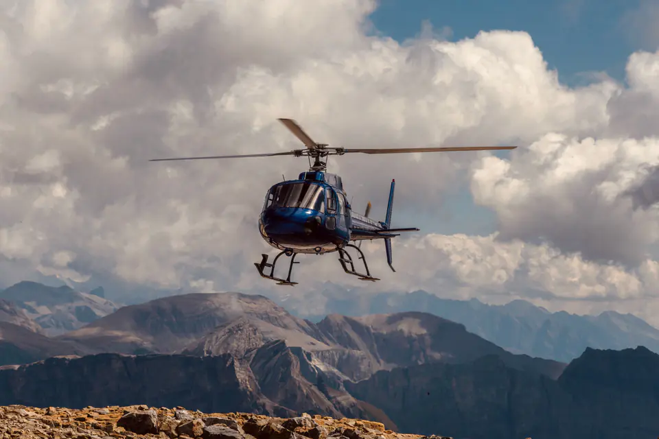 A dark blue helicopter flying over bare rocky mountains under towering clouds