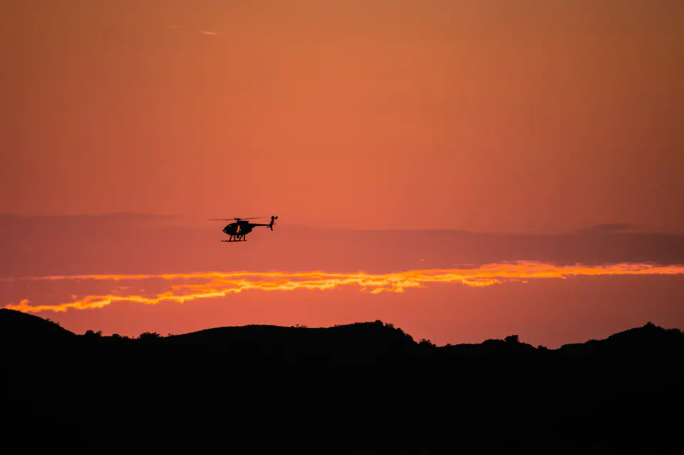 A helicopter in silhouette against an orange dusk sky above dark hills